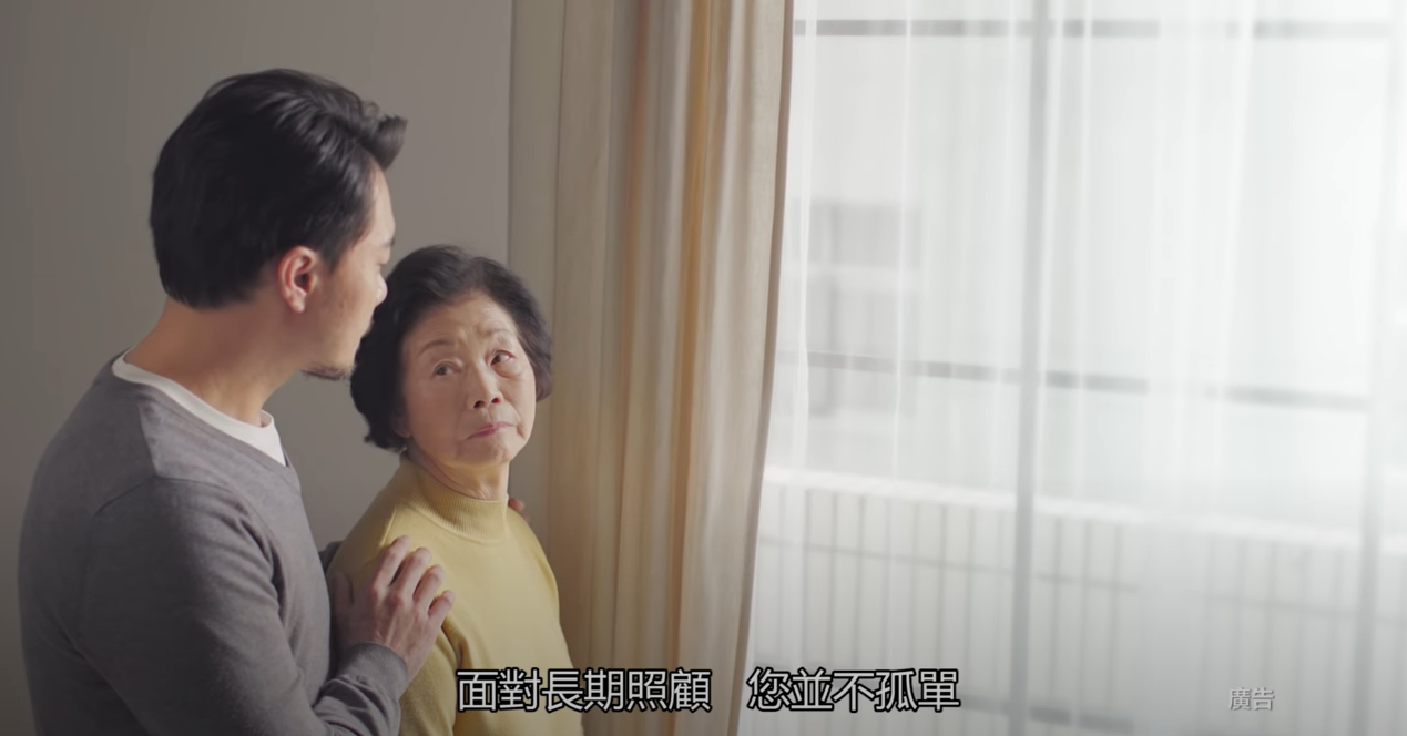 畫面中有一位長輩抬頭望著右側的家屬，下方字幕顯示「面對長期照顧，您並不孤單」